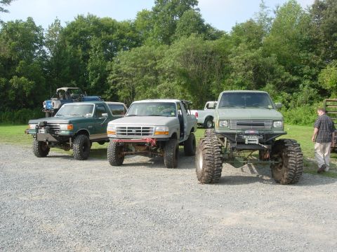 1995 Ford Bronco - Several modified trucks are parked on a gravel lot with trees in the background.