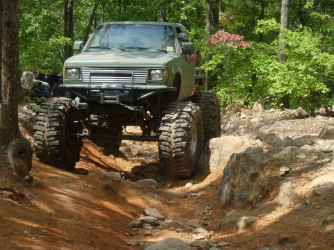 1995 Ford Bronco - Off-road truck with oversized tires climbing a challenging rocky path in a wooded area.