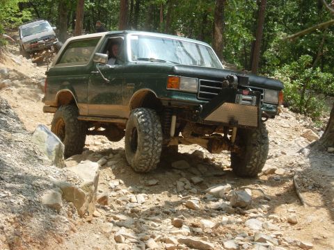 1995 Ford Bronco - Two off-road vehicles navigate a steep, rocky path through a wooded area.