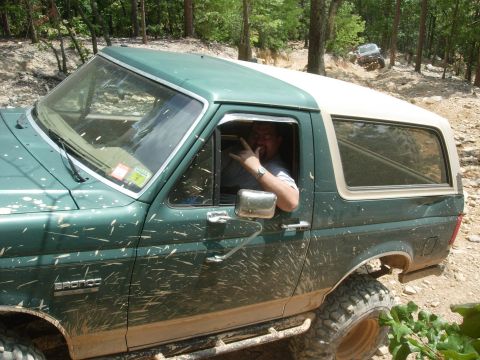 1995 Ford Bronco - A mud-covered green Ford Bronco with a white top navigates an off-road trail, with another vehicle visible in the background.