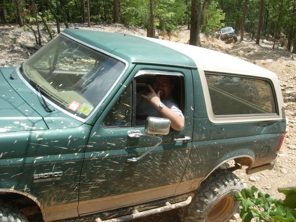 Green Ford Bronco with a white top, covered in mud, driving off-road