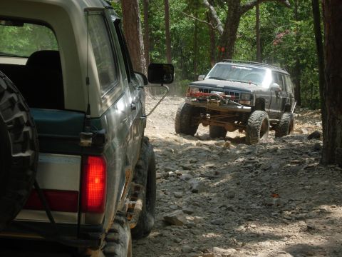1995 Ford Bronco - Two modified SUVs tackle a challenging, rocky off-road trail surrounded by trees.
