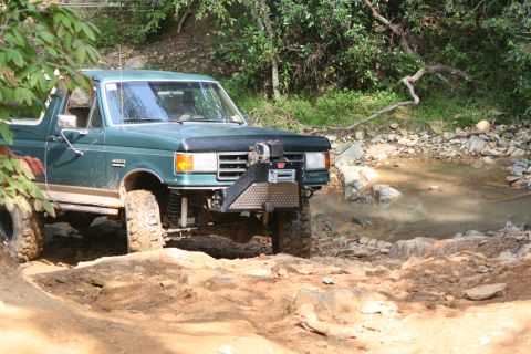 1990 Ford Bronco - 1990 Ford Bronco tackling an off-road trail with water and rocks.