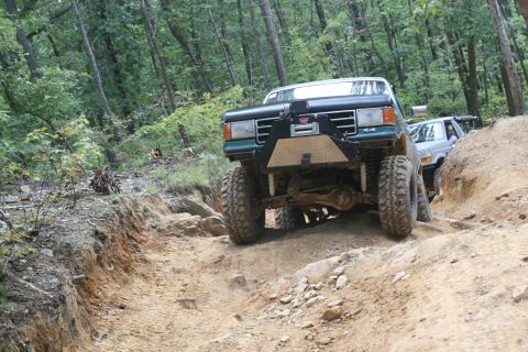 1990 Ford Bronco - Two SUVs navigate a challenging dirt path through a wooded area.