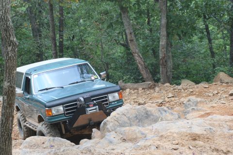1990 Ford Bronco - This Ford Bronco is tackling a challenging off-road course with large rocks and trees surrounding it.