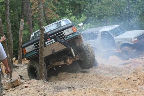 1990 Ford Bronco - Two off-road vehicles navigate a challenging rocky terrain in a wooded area.