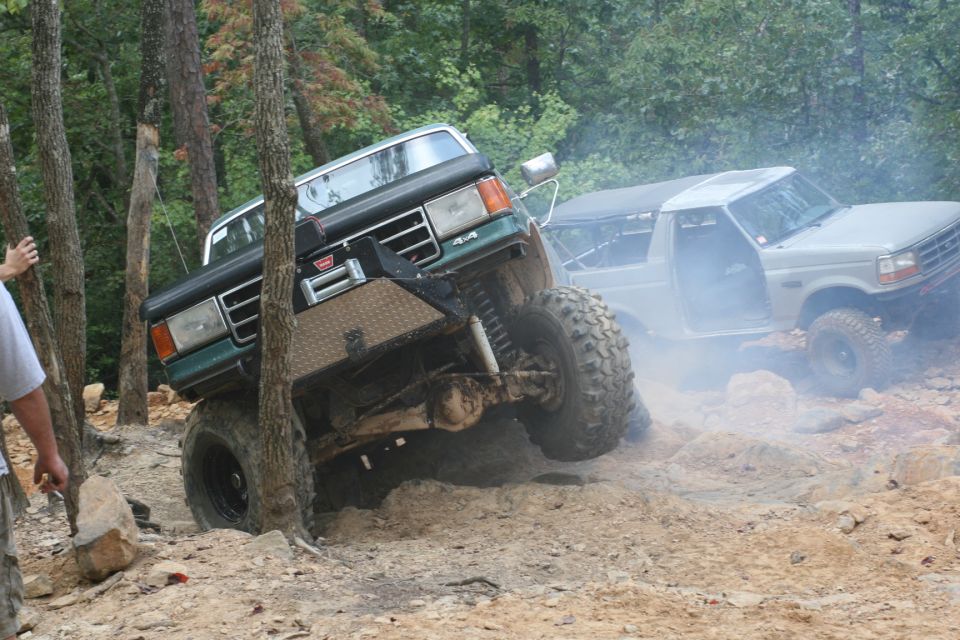 Gray Ford Bronco with doors removed, driving on a rocky, wooded trail with smoke around it