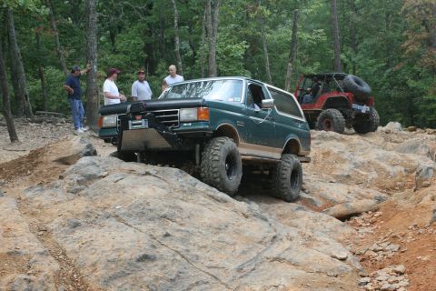 1990 Ford Bronco - Two off-road vehicles navigate a rocky trail with spectators observing.