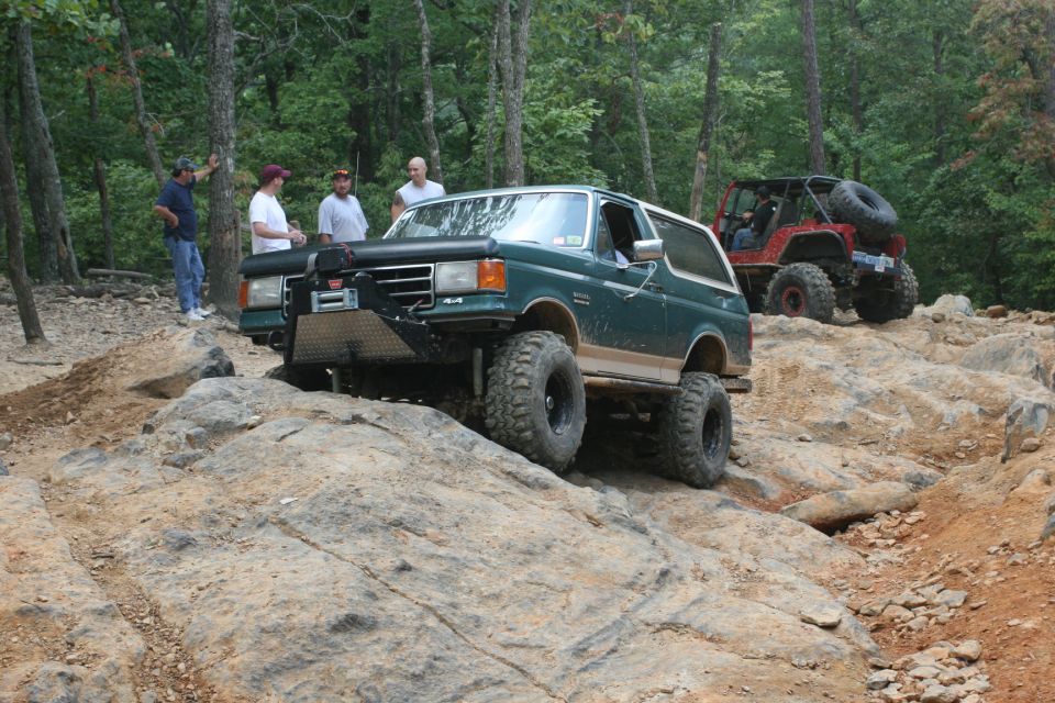 Green Ford Bronco climbing rocks, red Jeep behind it. People watching