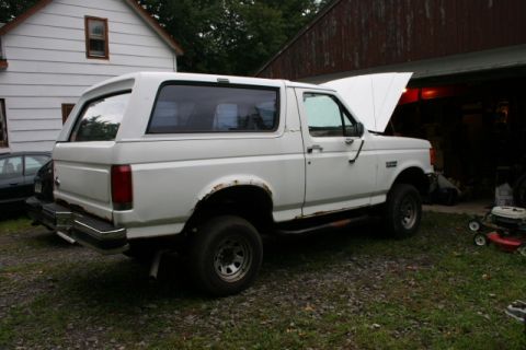 1987 Ford Bronco - This white 1987 Ford Bronco is seen parked outside a garage with its hood raised, revealing the engine bay.
