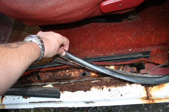 Hand lifting rubber weatherstrip to reveal rusted-through floor pan under red carpet at door sill