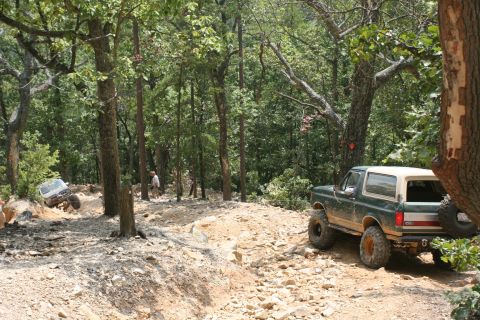 1990 Ford Bronco - Two off-road vehicles navigate a challenging rocky trail through a wooded area.
