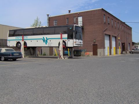1979 Ford Bronco - A large white bus is elevated on a lift in front of a brick building, with a dark sedan parked nearby.