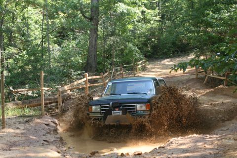 1990 Ford Bronco - 1990 Ford Bronco navigating a muddy trail with trees and a wooden fence.