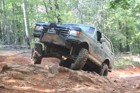 1990 Ford Bronco - Muddy dark green Ford Bronco climbing a red-clay and rock ledge in the woods, front driver tire lifted off the ground. It has a diamond-pla…