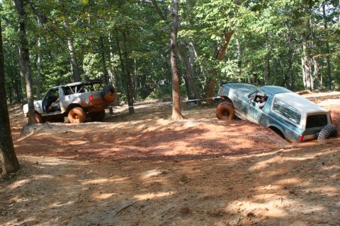 1990 Ford Bronco - Two off-road vehicles are deeply stuck in the mud on a trail surrounded by trees.