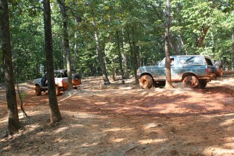 1990 Ford Bronco - Two off-road vehicles navigate a muddy trail through a wooded area.