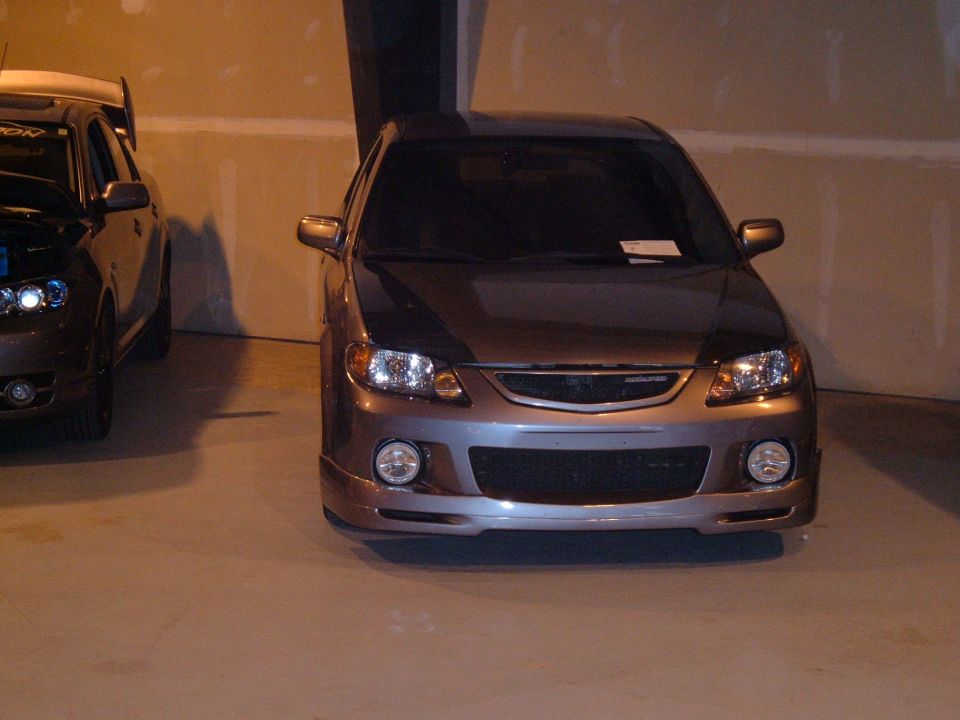 Front view of a modified car with aftermarket bumper and headlights. Another car is partially visible to the left