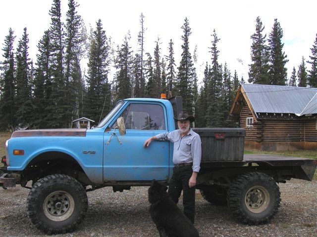 Man in cowboy hat and blue shirt leans on side of light blue pickup truck with flatbed and large tires. Black dog sits in front of truck. Log cabin and pine trees in background