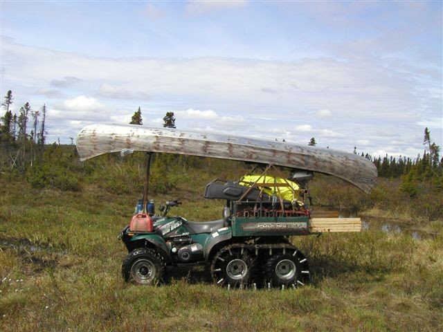 Green ATV with tracks, carrying a canoe and lumber in a grassy, wooded area