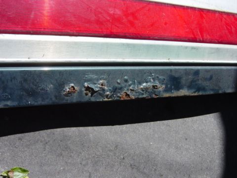 1991 Ford Bronco - The rear bumper corner of a dark green vehicle displays significant rust, with a portion of the red taillight visible above.