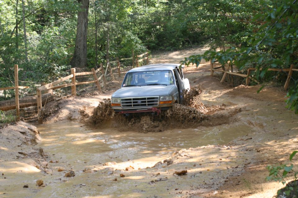 Gray Ford Bronco splashing through a muddy trail with wooden fences on either side