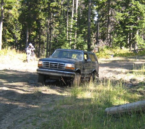 1995 Ford Bronco - A dark SUV navigates a dirt path in a wooded area, with a dirt bike rider visible in the distance.