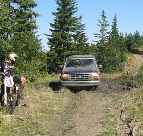 1995 Ford Bronco - A black 1995 Ford Bronco navigates a muddy trail surrounded by trees, with a dirt road winding uphill in the background.