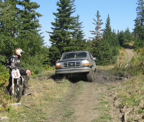 1995 Ford Bronco - Ford Bronco navigates a challenging muddy off-road path while a dirt bike rider observes.