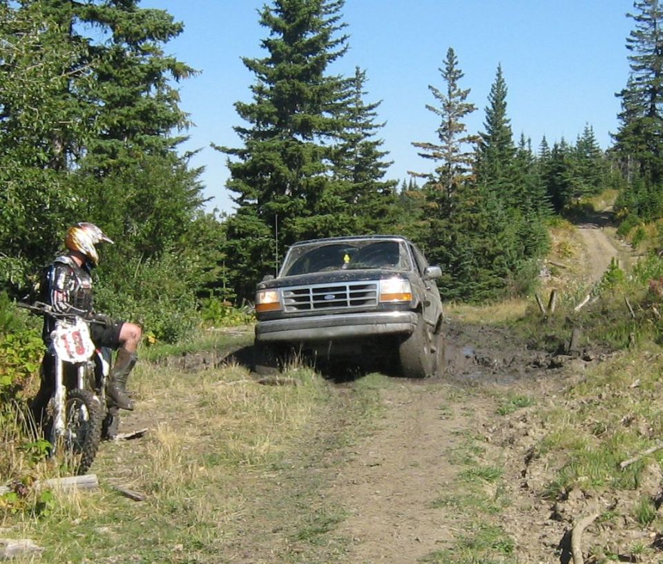 1995 Ford Bronco driving through a muddy trail with a dirt bike rider watching