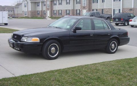 2003 Ford Crown Victoria - Side profile of a black 2003 Ford Crown Victoria sedan parked in front of apartment buildings.