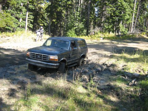 1995 Ford Bronco - A dark blue 1995 Ford Bronco navigates a muddy trail through a forest, with a dirt bike rider visible in the background.
