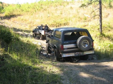 1995 Ford Bronco - Two off-road vehicles navigate a muddy trail through a wooded area.