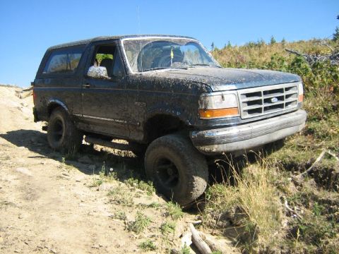 1995 Ford Bronco - A muddy Ford Bronco navigates an off-road dirt path under a clear blue sky.
