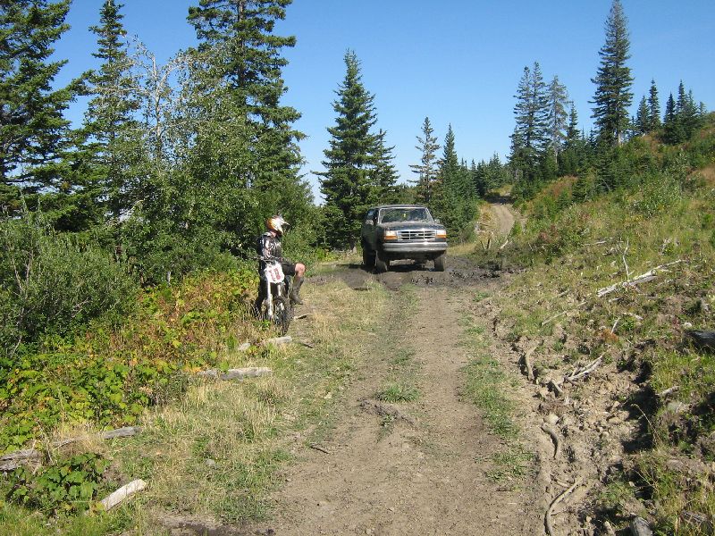 Ford Bronco driving on a muddy dirt road with trees and a dirt bike on the side