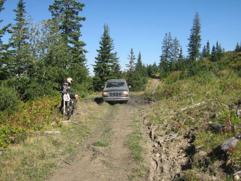1995 Ford Bronco - A dirt bike rider watches a Ford Bronco navigate a muddy trail through a wooded area.