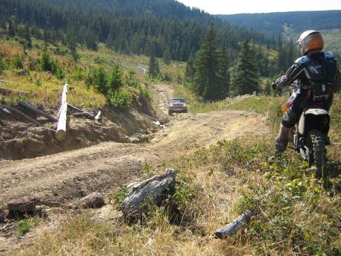 1995 Ford Bronco - Dirt bike rider watches an SUV navigate a challenging dirt road in a mountainous, forested area.