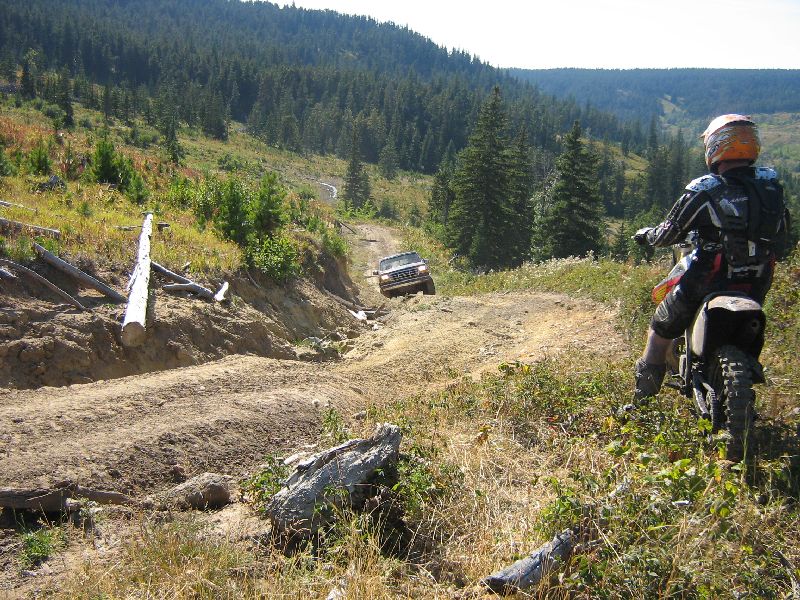 Dirt bike rider looks at SUV driving up a steep, rutted dirt road in a wooded, hilly area