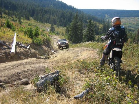 1995 Ford Bronco - A dirt biker observes a white Ford Bronco navigating an off-road trail through a wooded mountainside.