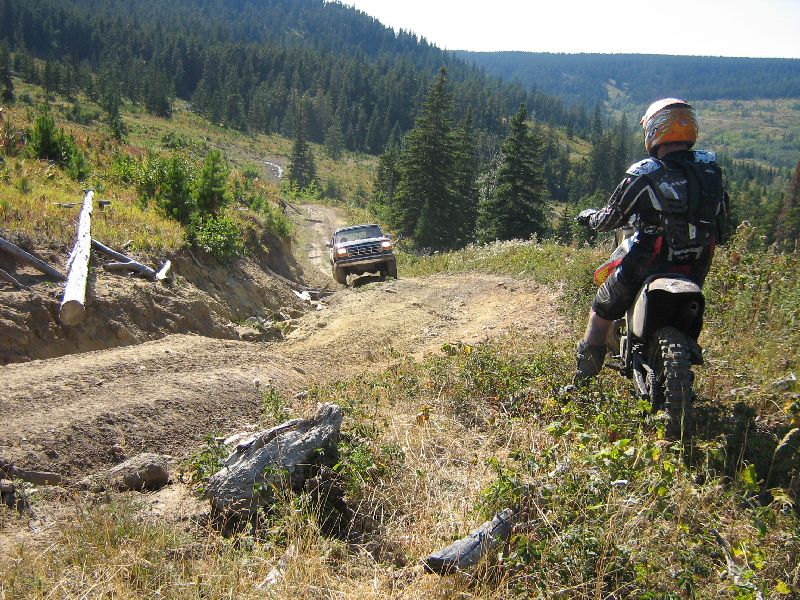 White Ford Bronco driving up a dirt road in a forest, with a dirt biker in the foreground