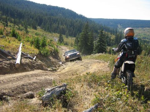 1995 Ford Bronco - A white SUV navigates a challenging dirt road while a dirt bike rider observes from the side.
