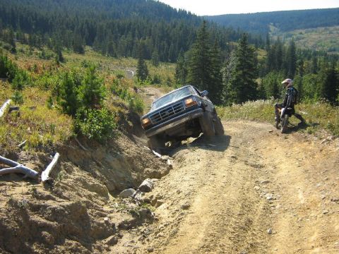 1995 Ford Bronco - A dark SUV navigates a challenging, rutted dirt path while a dirt bike rider observes the difficult terrain.