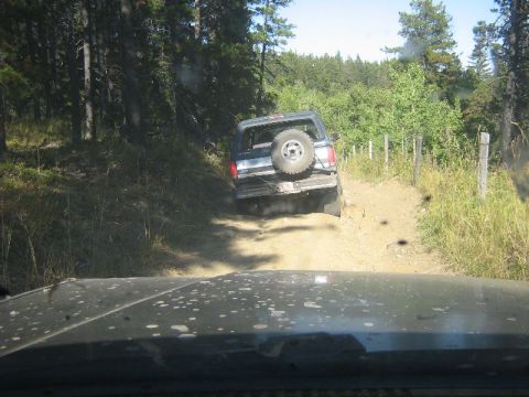1995 Ford Bronco - A dark SUV with a spare tire mounted on its rear door navigates a dirt road, followed by another vehicle.