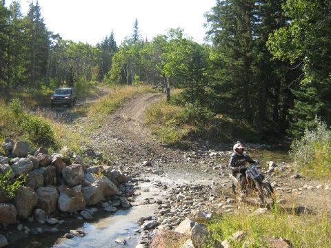1995 Ford Bronco - Two vehicles navigate a rough, wooded terrain with a stream crossing.