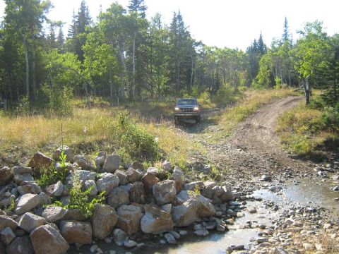 1995 Ford Bronco - A red Ford Bronco navigates a rocky dirt path and stream in a wooded area.