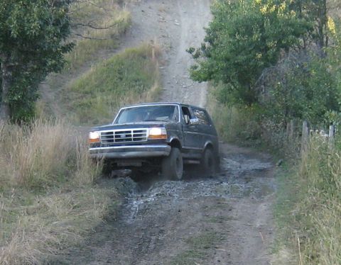 1995 Ford Bronco - A dark 1995 Ford Bronco navigates a challenging muddy terrain on a dirt road.