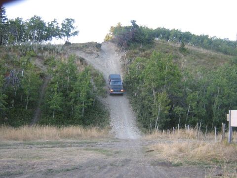 1995 Ford Bronco - A dark 1995 Ford Bronco navigates a steep dirt incline surrounded by trees and dry grass.