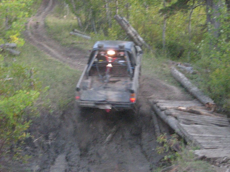 Pickup truck driving on a muddy, wooded trail, crossing a makeshift wooden bridge