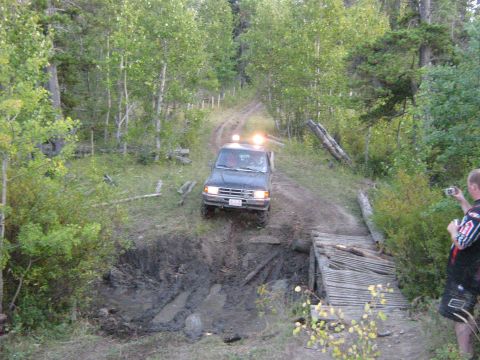 1995 Ford Bronco - A dark SUV navigates a challenging off-road path with a muddy obstacle and a rickety wooden bridge.