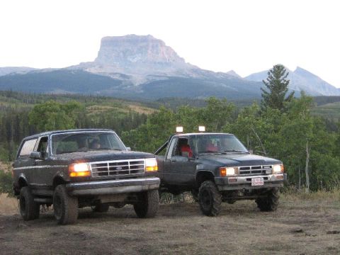 1995 Ford Bronco - Two off-road vehicles, a Ford Bronco and a Toyota pickup, are parked on a dirt path with a mountainous landscape behind them.